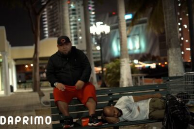 Dos hombres en un banco nocturno junto a un carrito de compras y edificios iluminados 6PzT0l6AxIg.