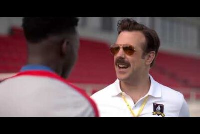 Entrenador sonriente con bigote y gafas de sol conversando en el campo 8PmX7zEUg w.