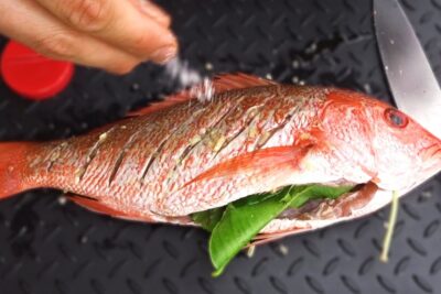 Mano espolvoreando sal sobre un pargo rojo entero con cortes en la piel y relleno de hojas verdes IIYUyi2 sNU