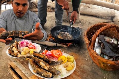 Persona mostrando pescado frito con platos de arroz, papas y cebolla junto a sartén al fuego y canasto de pescados ISBQJoKYOn4