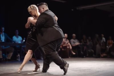 Pareja bailando tango en abrazo cerrado sobre un escenario con público observando LRVKkyW7n48