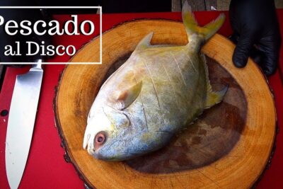 Pescado entero colocado sobre una tabla redonda de madera junto a un cuchillo y una mano con guante negro