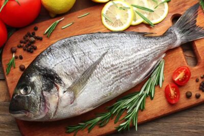 Pescado entero sobre tabla de madera con rodajas de limón, tomates cherry, romero y granos de pimienta bv0O5FcfPhY