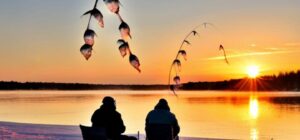 Dos pescadores sentados en sillas sobre la nieve junto a un lago helado al atardecer con cañas y peces colgando, carnada ideal pesca hielo consejos sorprendentes