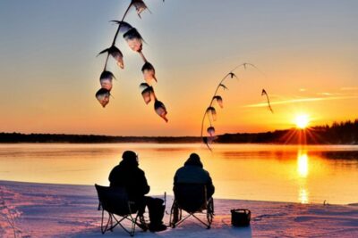 Dos pescadores sentados en sillas sobre la nieve junto a un lago helado al atardecer con cañas y peces colgando, carnada ideal pesca hielo consejos sorprendentes