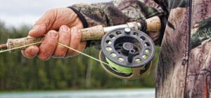 Mano sosteniendo una caña y carrete de pesca con línea verde, imagen útil para consejos comprar linea pesca mosca agua salada.