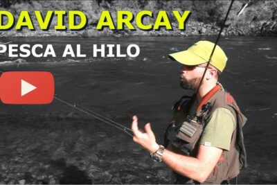 Pescador con gorra y chaleco practicando pesca al hilo junto a un río, con overlay de reproductor y texto iXjXXsUVsTA.