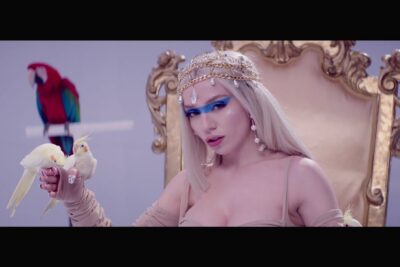 Mujer rubia con maquillaje azul, tocado de cadenas y aves exóticas en un trono jH1RNk8954Q