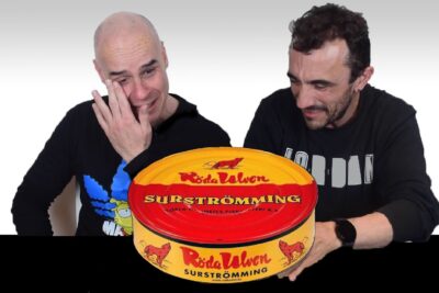 Dos hombres reaccionan sorprendidos ante una lata de surströmming kPnzcLdnMBE con diseño rojo y amarillo