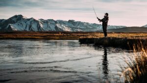 Pescador en la orilla lanzando la caña frente a un río y montañas nevadas metodos ilegales pesca conoce tecnicas pescadores