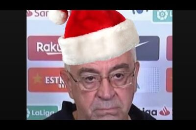 Hombre mayor serio con gorro de Santa frente a un panel de prensa pdJuLkUQa54
